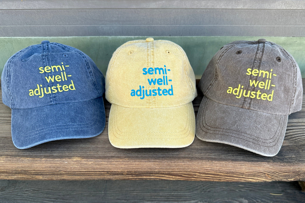 Three baseball caps with 'semi-well-adjusted' text on a wooden surface.