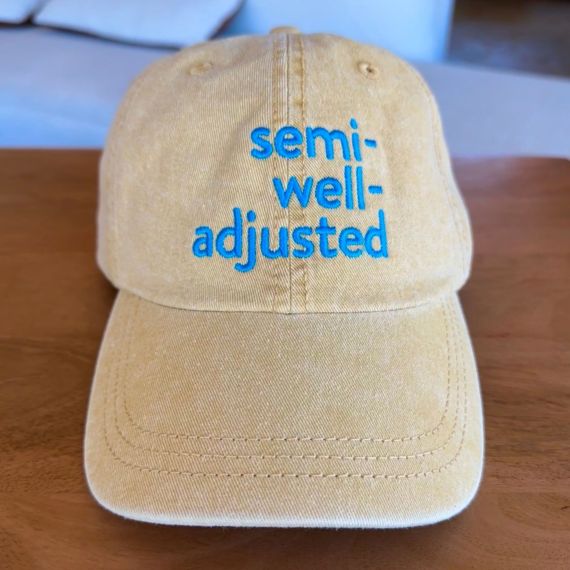 Butter yellow cap with 'semi-well-adjusted' text in blue on a wooden surface.